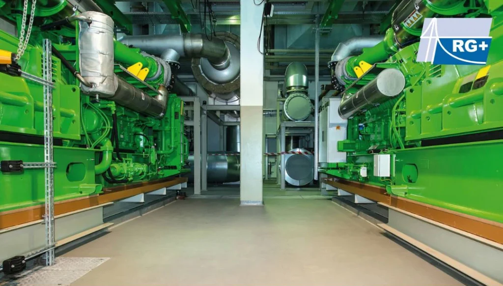 Zwei grüne Generatoren in einem technischen Raum mit Rohren und Lüftungssystemen.