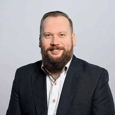 Dominik Bartz, ein Mann mit Bart, trägt einen Anzug und lächelt in die Kamera.