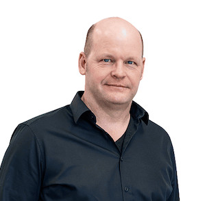 Christian Kahmen in einem schwarzen Hemd mit kurzem Kragen, lächelnd vor einem weißen Hintergrund.