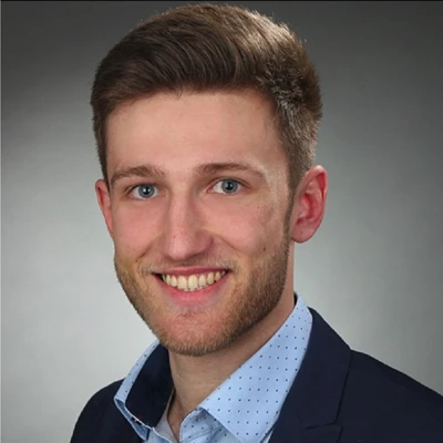 Paul Weiser lächelt in einem professionellen Porträt mit Anzug und Hemd mit Punkten.