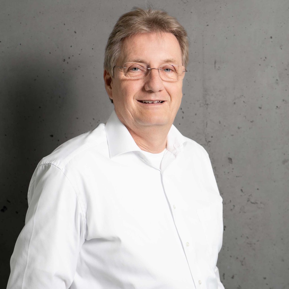 Roman Mielinski in einem weißen Hemd vor einer grauen Wand mit neutralem Hintergrund.