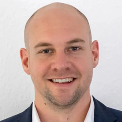 Stefan Nitz mit Glatze und Anzug, lächelt in die Kamera vor einem weißen Hintergrund.