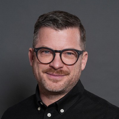 Sven Werdehausen mit Brille und Bart, lächelnd vor einem grauen Hintergrund.