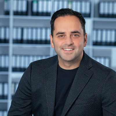 Ersan Tekan in einem schwarzen Blazer mit einem lächelnden Gesichtsausdruck vor einem Regal mit Aktenordnern.