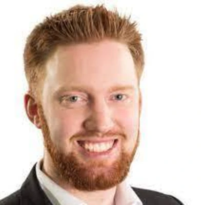 Tom Frenzel mit rotem Bart und lächelndem Gesicht, trägt einen Anzug mit Hemd und Krawatte.