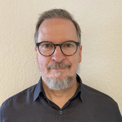 Uwe Ulrich mit Brille und Bart, in einem Hemd mit Kragen, vor einer hellen Wand.
