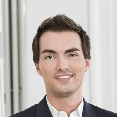 Arne Thorben Damm mit einem freundlichen Lächeln in einem formellen Anzug vor neutralem Hintergrund.