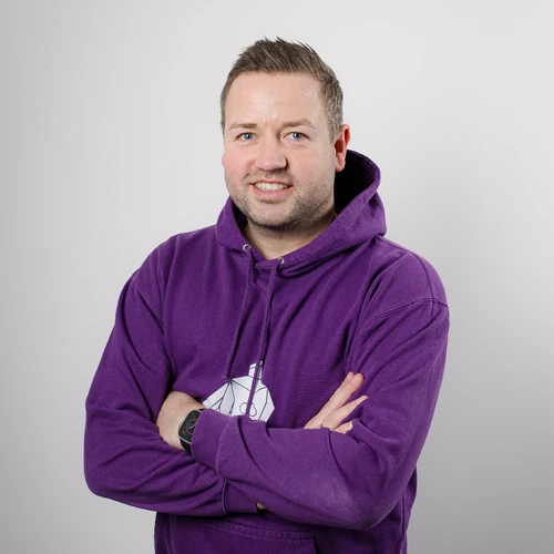 Nils Kathagen trägt einen lila Hoodie und steht mit verschränkten Armen vor einem neutralen Hintergrund.