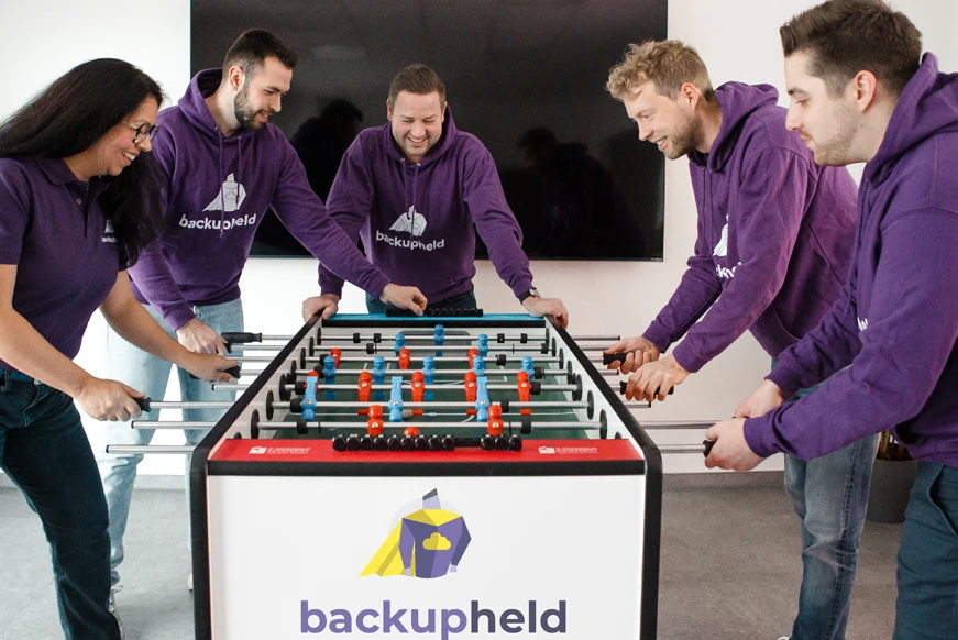 Fünf Personen spielen Tischfußball an einem Tisch mit dem Logo von backupheld GmbH.