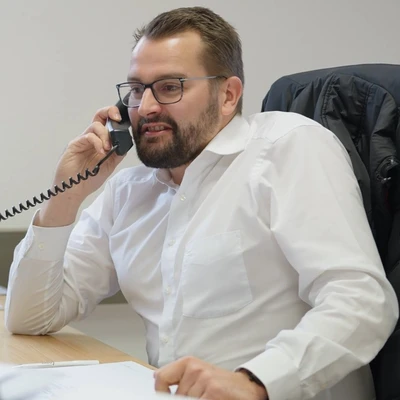 Jonas Knepper am Telefon, in einem Büro mit einem weißen Hemd und Brille, sitzt an einem Tisch.