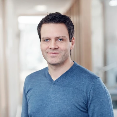 Mann mit blauer Pullover steht in einem modernen Büro mit neutralem Hintergrund.