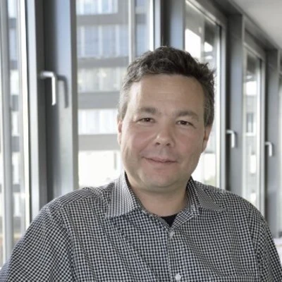 Leonhardt, ein Mann mit einem karierten Hemd, sitzt vor großen Fenstern in einem modernen Raum.