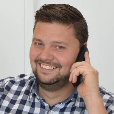 Nico Hoeller lächelt und spricht am Telefon, trägt ein kariertes Hemd.