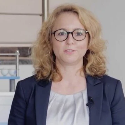 Ruschke, eine Frau mit lockigem, blondem Haar und Brille, trägt einen blauen Blazer und steht vor einem hellen Hintergrund.